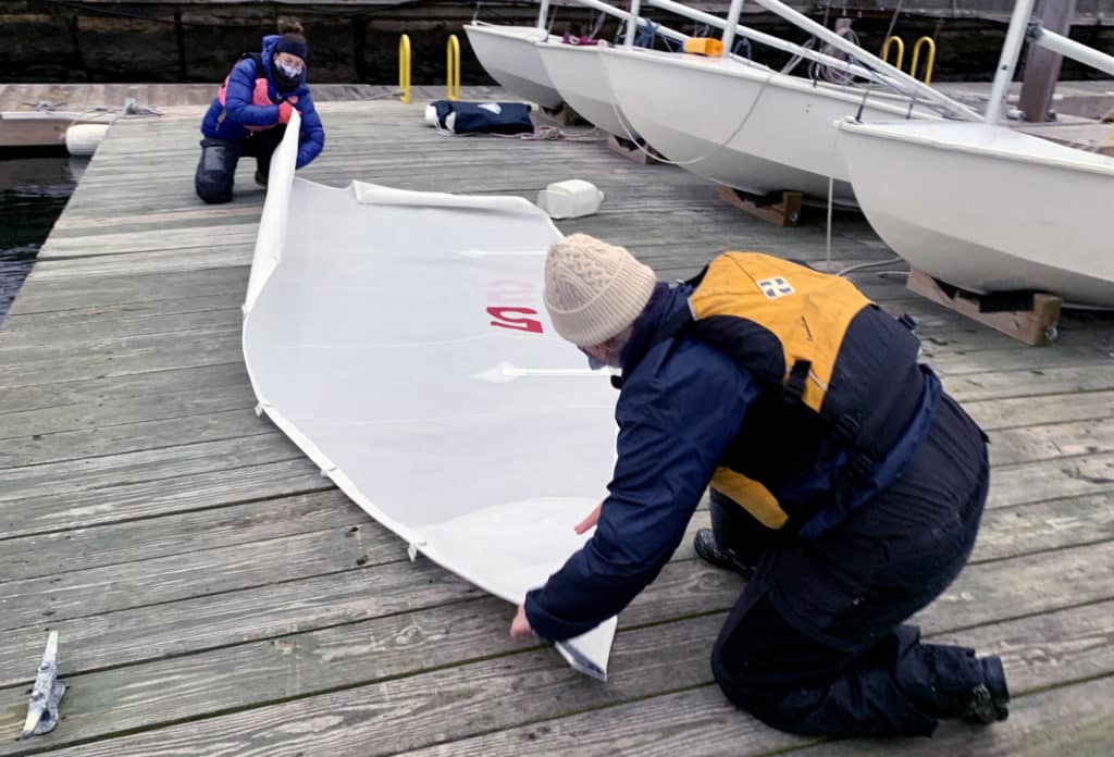 two sailors in foul weather gear and lifejackets fold a sail on the dock